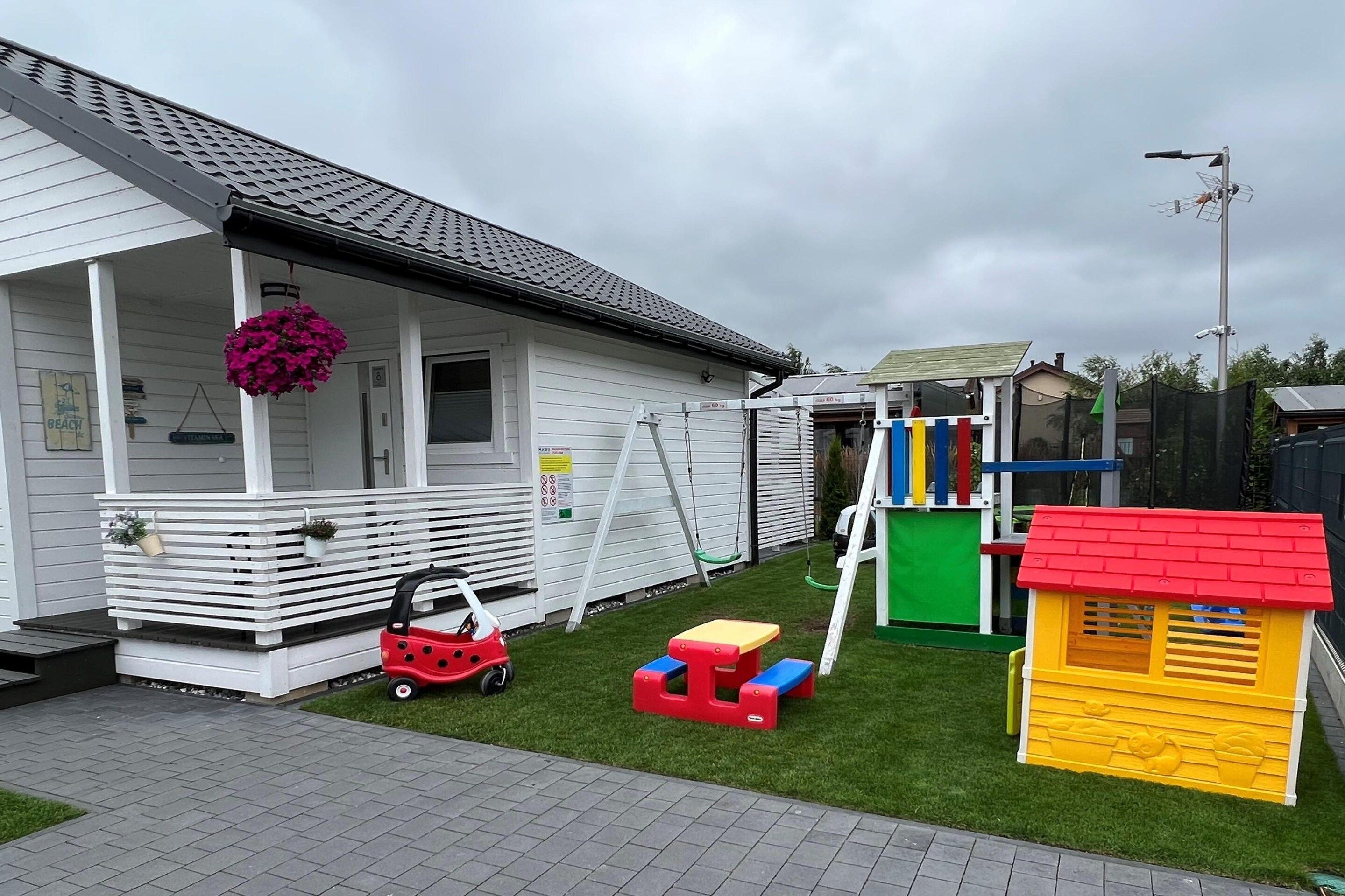 A white building with a porch, a swing set, a small red toy car, and a yellow playhouse.