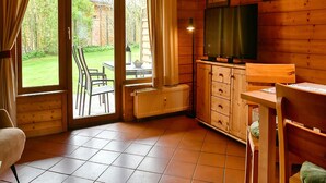 Uma cabana acolhedora com paredes de madeira, uma área de refeições com mesa e cadeiras, uma televisão e vista para um jardim pela porta de vidro deslizante.