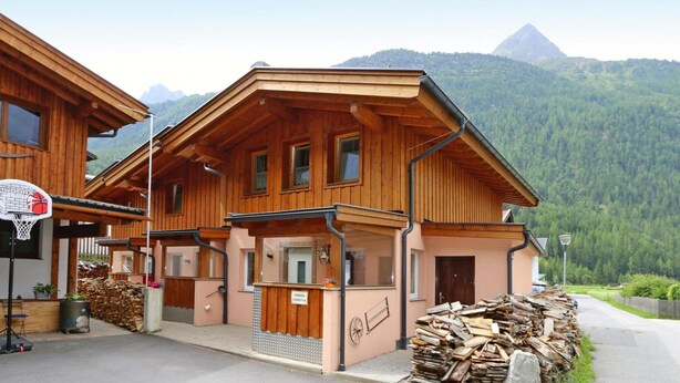 A wooden chalet with a basketball hoop, a small outdoor seating area, and a view of a mountain.