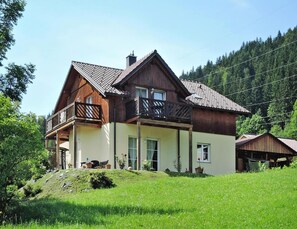 Una casa de madera de dos pisos con un balcón, rodeada de un paisaje verde exuberante y una colina boscosa al fondo.