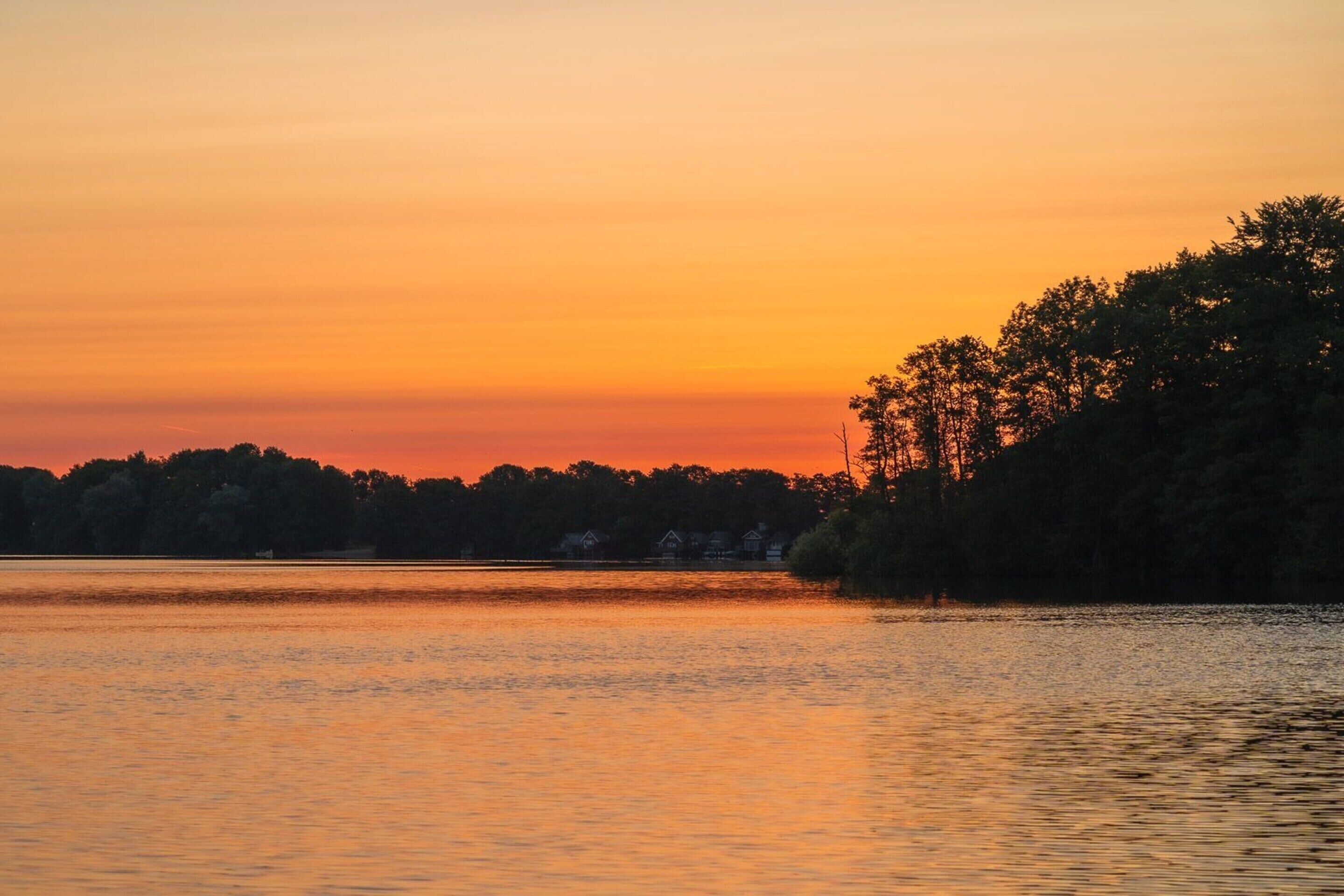 A sunset over a calm lake with a forested shoreline and houses in the background.