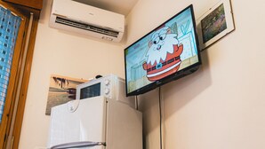 A room with a wall-mounted TV displaying a cartoon character, a white refrigerator, and an air conditioning unit. - Condominio Quercia in Bibione Lido del Sole (San Michele al Tagliamento)
