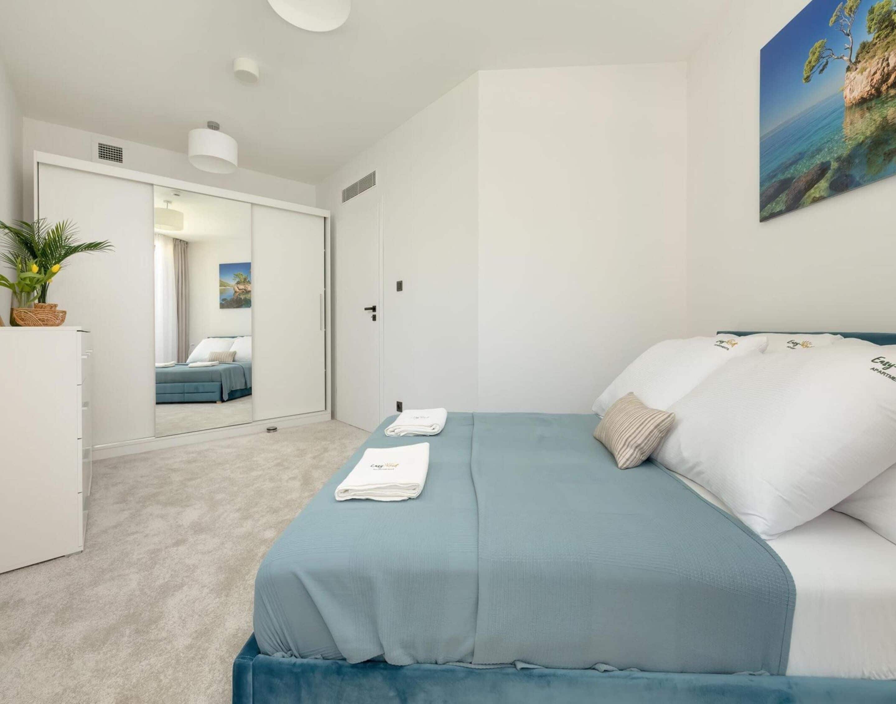 A modern bedroom with a bed, a dresser, a mirror, and a painting of a coastal scene.
