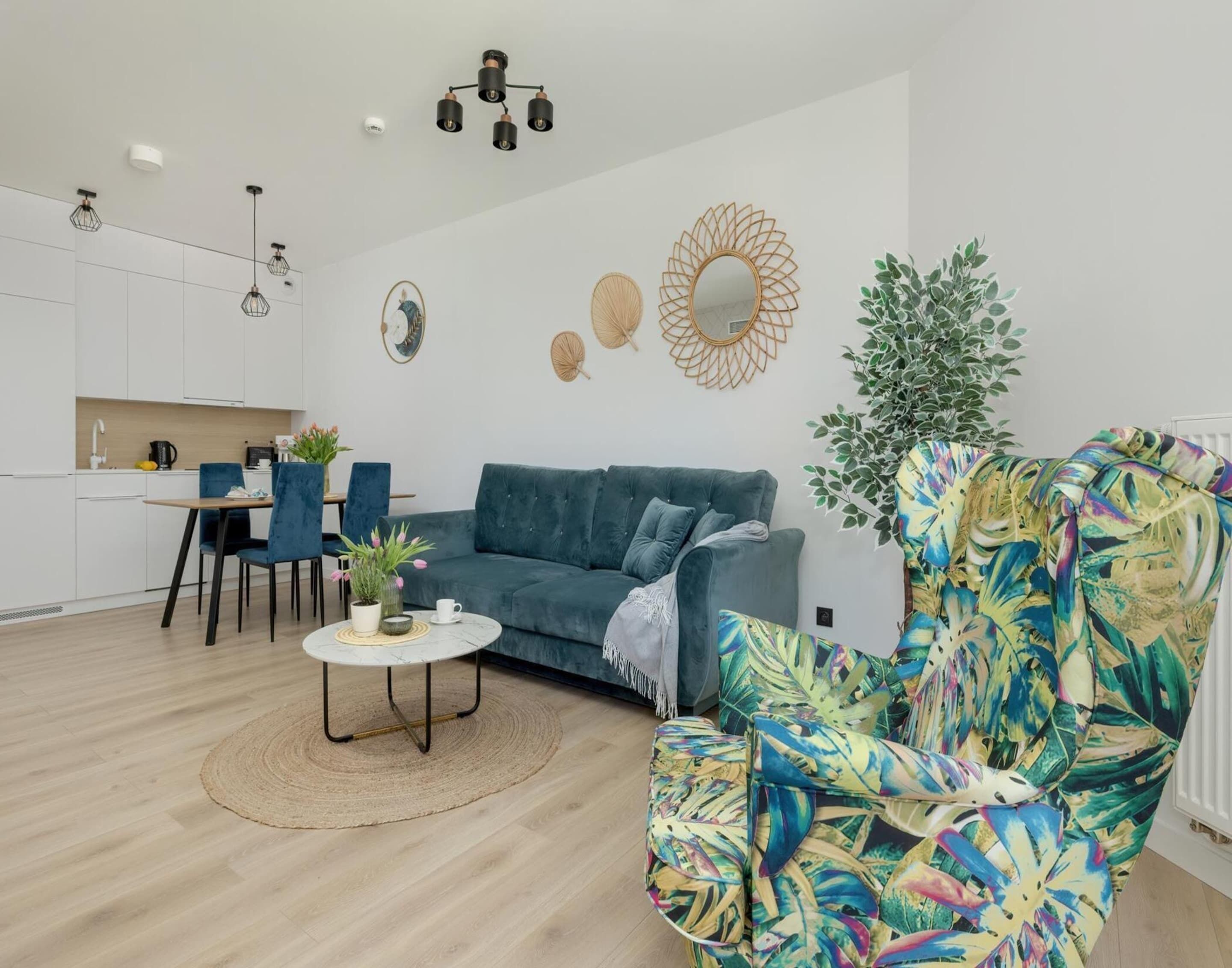 A modern living room with a teal sofa, a vibrant floral armchair, a small coffee table, and a kitchen in the background.