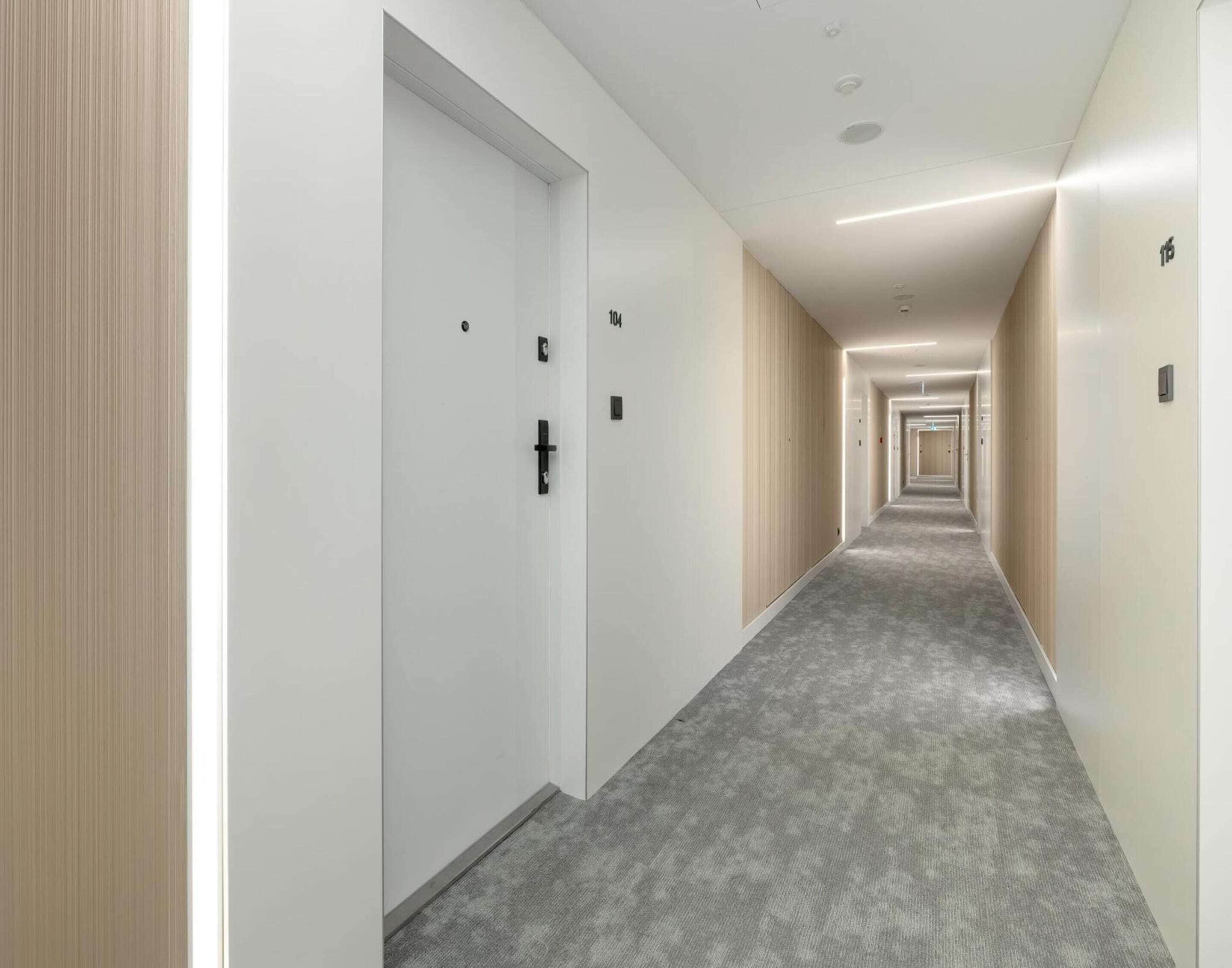 A long, narrow hotel corridor with doors on both sides, carpeted floors, and recessed lighting.
