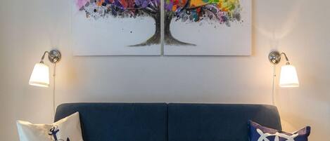 A room with a blue sofa, two wall-mounted lamps, and a colorful abstract painting above the sofa.