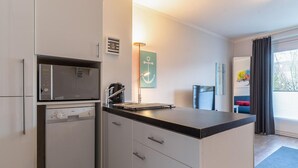 Een compacte keuken met een magnetron, koffiezetapparaat en een klein spoelgedeelte. In de aangrenzende woonkamer staan een stoel en een bank.