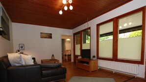 A living room with a black leather sofa, a wooden entertainment unit, and a large window with blinds. - Ferienhaus in der Nahe des Sees, Haustierfrei (Clausthal-Zellerfeld)