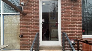 A brick building with a glass door entrance, a metal staircase with handrails, and a small outdoor seating area.