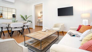 A modern living room with a wooden coffee table, white sofas, a dining area with chairs, a TV mounted on the wall, and a kitchen visible through an open door.
