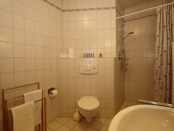 A bathroom with a toilet, a shower, and a bathtub. There are towels hanging on a rack, a toilet paper holder, and a shelf with toiletries. - Reihenhauser in Strackholt (Grosefehn)