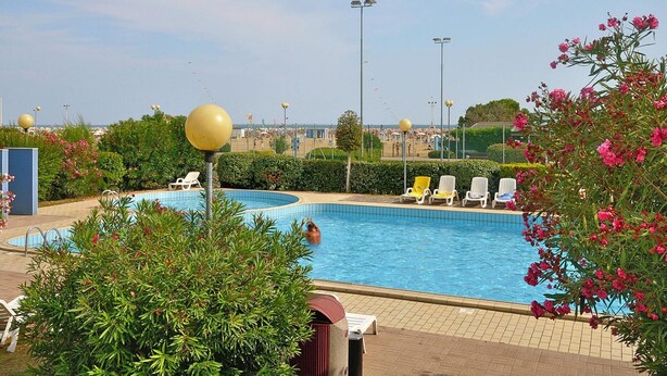 Ein Schwimmbecken mit Liegestühlen, einem Mülleimer und Blick auf Strand und Stadtbild.