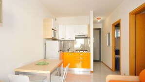 Eine kompakte Küche mit weißen Schränken, einem Herd und einer Mikrowelle. Im Wohnbereich befindet sich ein Esstisch mit zwei Stühlen und ein kleines orangefarbenes Sofa.
