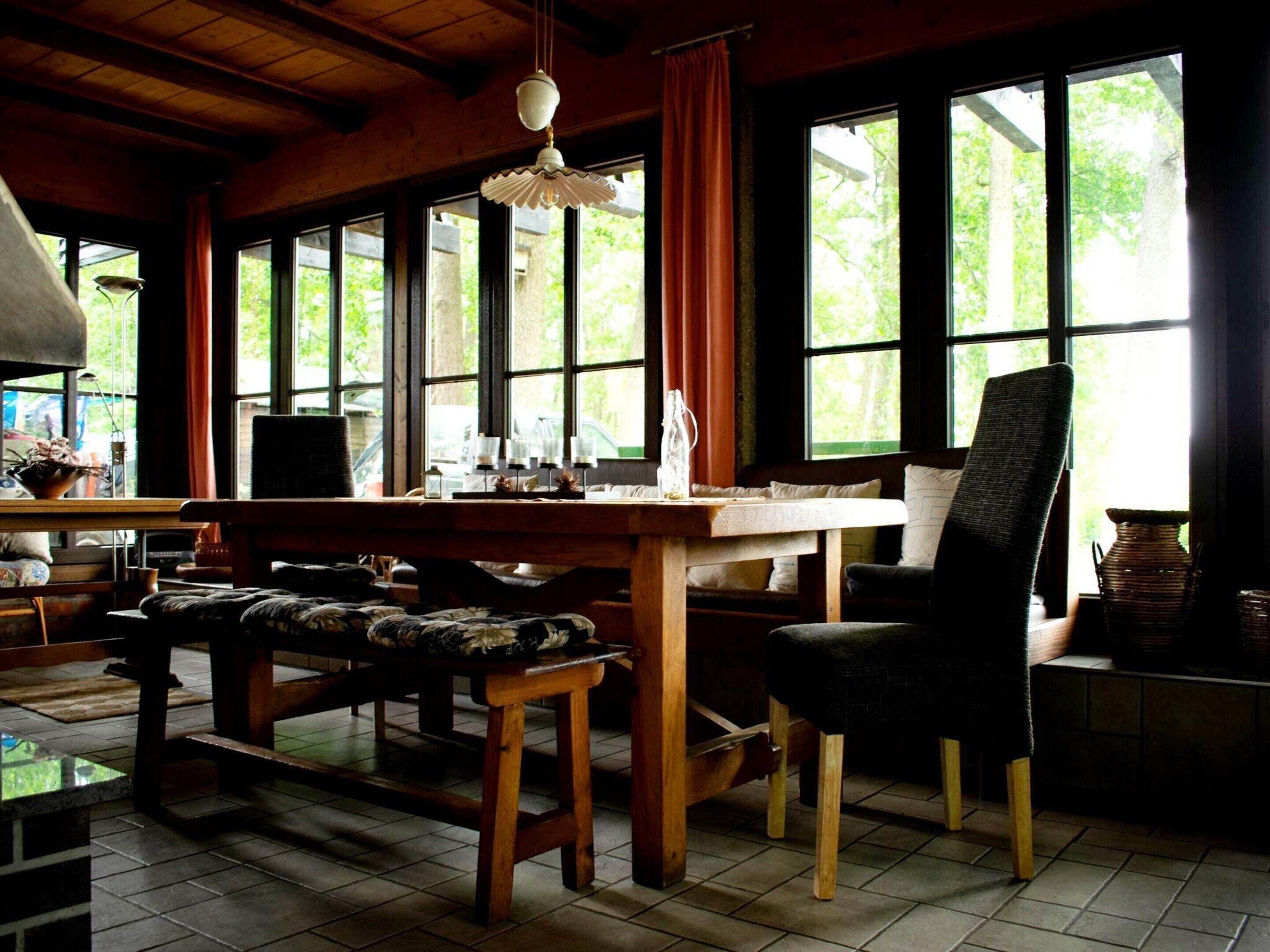 A cozy room with a wooden table, a bench, and a chair. Large windows allow natural light and views of greenery outside.