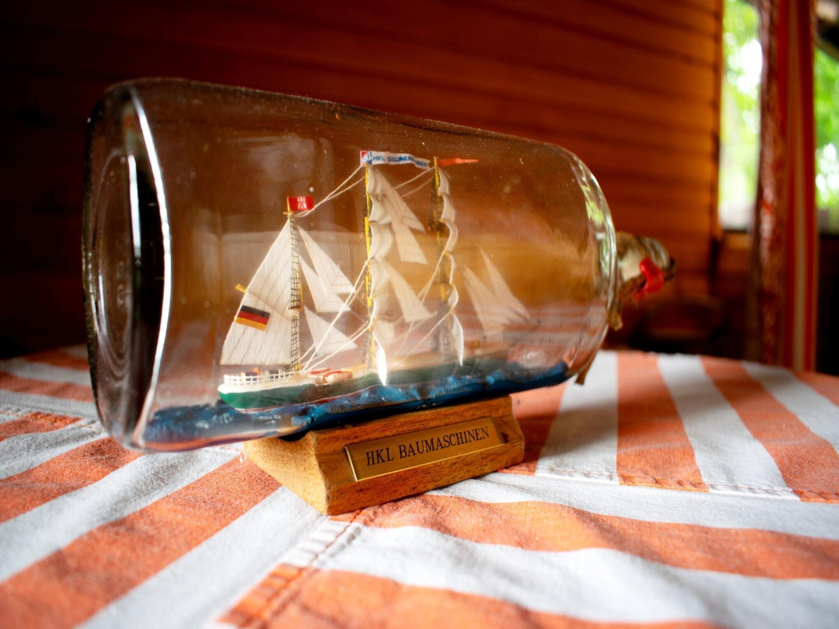 Een model van een schip in een glazen fles op een houten voetstuk, met een label waarop 