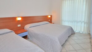 A hotel room with two beds, a wooden headboard, a nightstand, and a window with curtains. - Apartment in Bibione Near Beach and Pool (San Michele al Tagliamento)