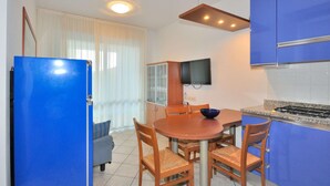 A compact kitchen with blue cabinets, a wooden dining table, and a TV mounted above it. - Apartment in Bibione Near Beach and Pool (San Michele al Tagliamento)