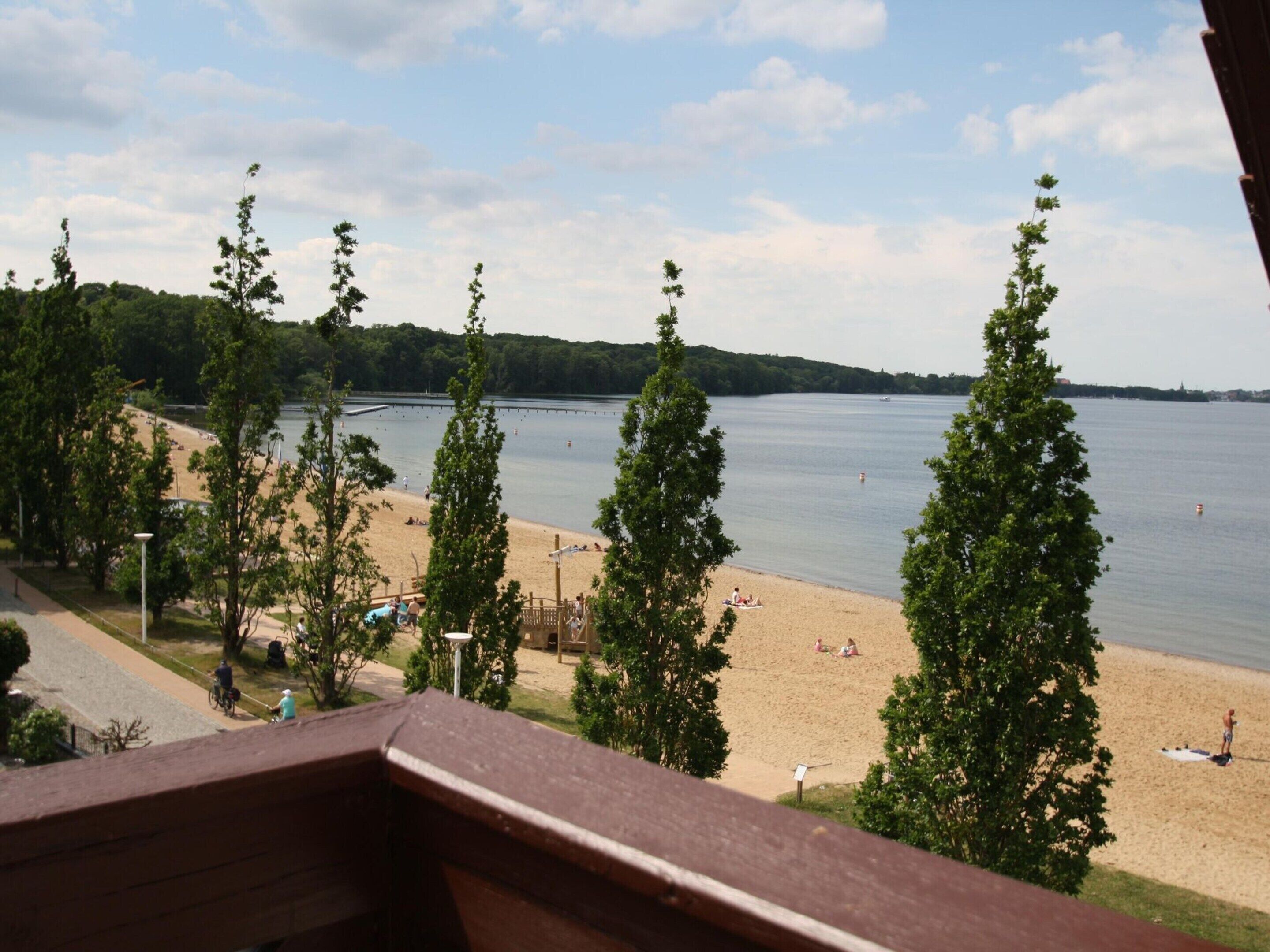 A beach view from a balcony with a wooden railing, trees, and a sandy beach with people and a lifeguard tower in the distance.