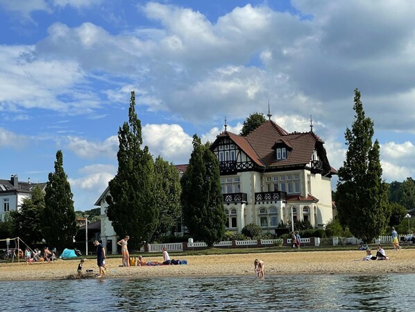A large building with a traditional architectural style, a sandy beach, and a body of water. - Ferienwohnung Am Strand in Schwerin (Schwerin)