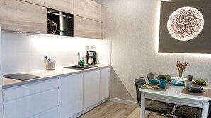 Moderni keittiö, jossa on valkoiset kaapit, sisäänrakennettu uuni, kahvinkeitin sekä ruokailutila, jossa on pöytä ja tuolit.