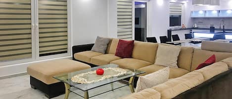 Un salon moderne avec un ensemble de canapés, une table basse en verre et un coin cuisine à l’arrière-plan.