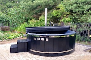A black hot tub with a metallic pole in the center, surrounded by a paved area with a fence and greenery in the background. - Holiday Home With Summer Pool + Sauna, Whirlpool (gmina Sianów)
