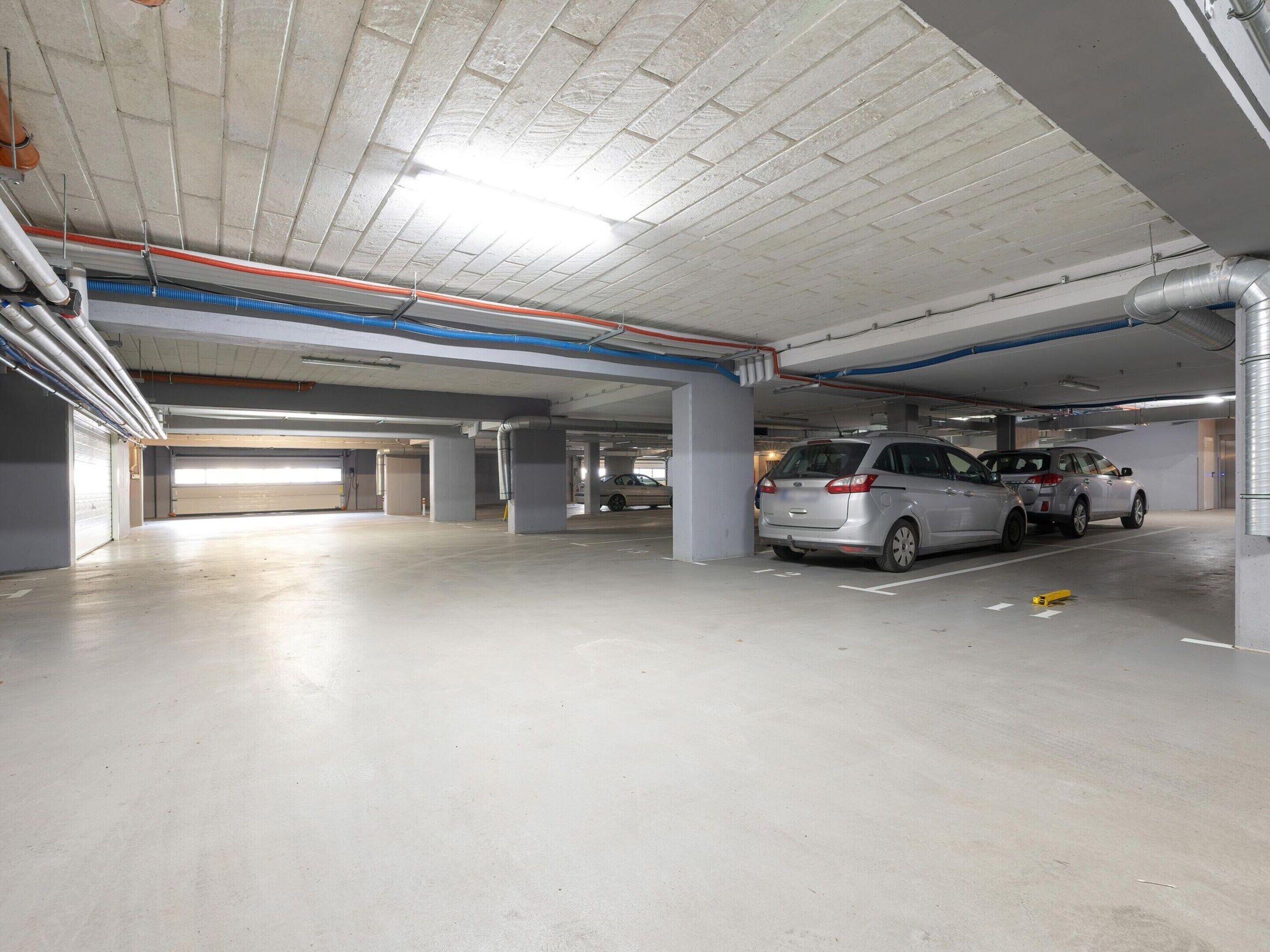 A multi-level parking garage with concrete floors and columns, featuring several parked cars and visible pipes and ducts on the ceiling.