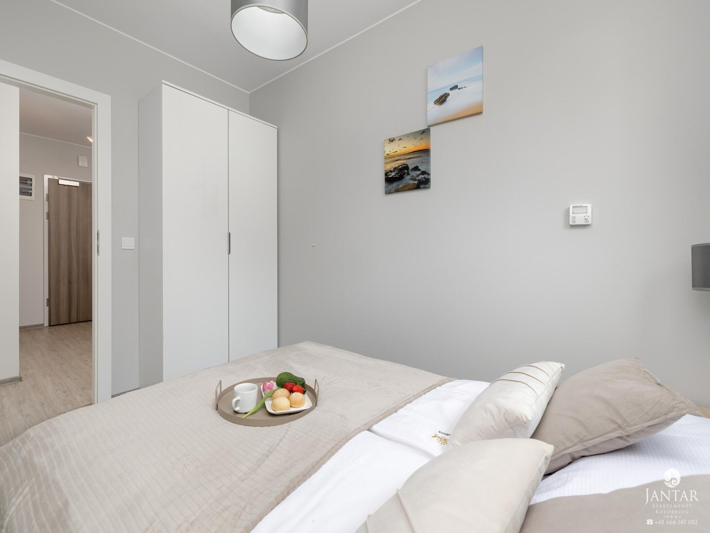 A modern bedroom with a bed, a tray with fruits and a cup, a thermostat, and a view of the sea through the window.