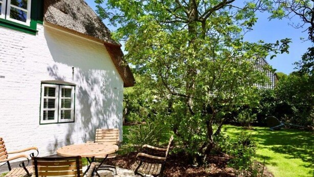 A white house with a thatched roof, a wooden table and chairs, and a tree in the background. - Reetgedecktes Ferienhaus in Nieblum auf Foehr (Nieblum)