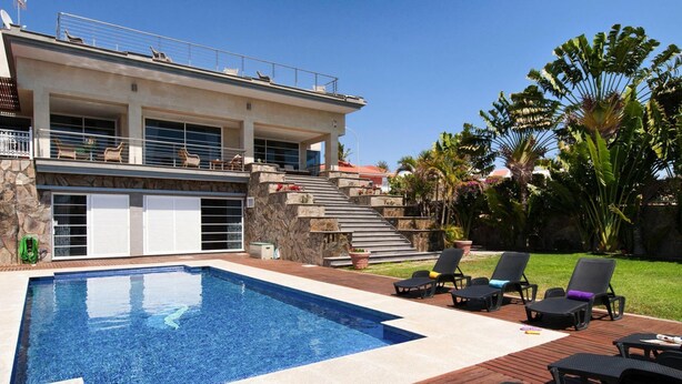 A swimming pool with a stone deck and lounge chairs, surrounded by a landscaped garden and a two-story building with a balcony. - Holiday Home, Maspalomas (San Bartolomé de Tirajana)