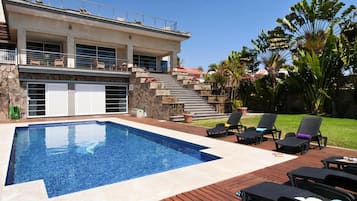 Piscina com deck de pedra e espreguiçadeiras, cercada por um jardim paisagístico e um edifício de dois andares com varanda.
