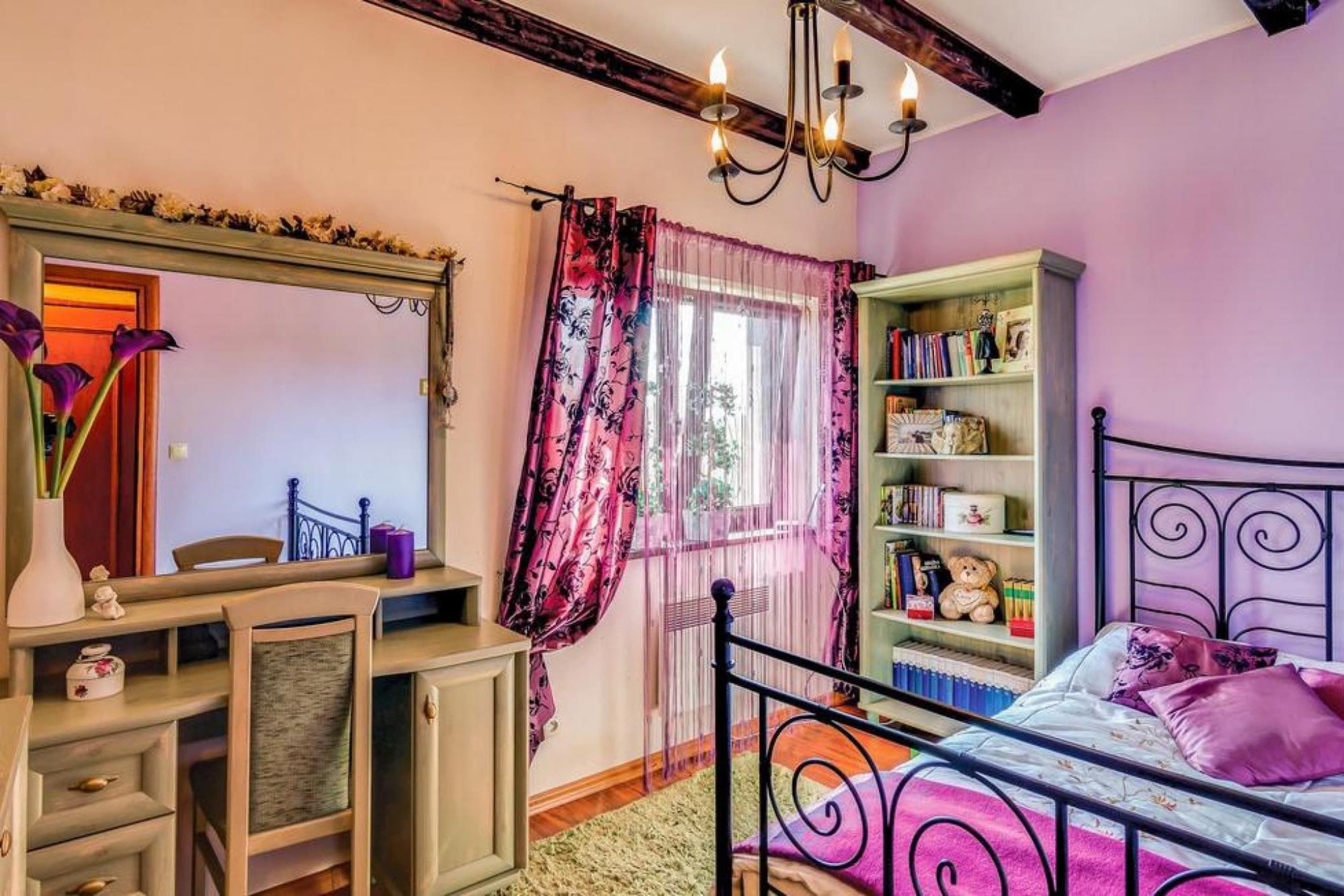 A bedroom with a bed, a bedside table, a dresser, a mirror, a window with curtains, and a bookshelf.