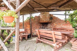 Ein Backofen aus Ziegelsteinen im Freien, unter einem hölzernen Vordach, mit Holzscheiten und einer Bank in der Nähe.