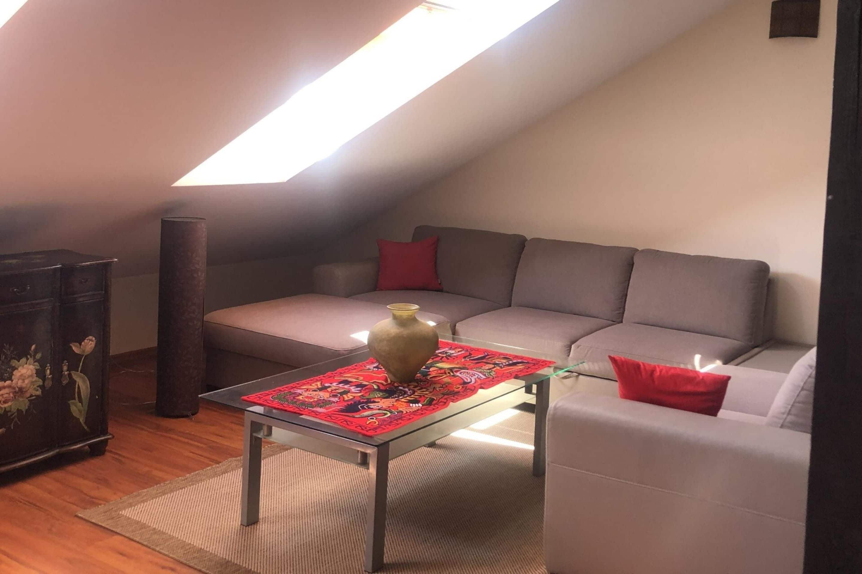 A cozy loft bedroom with a sloped ceiling, a grey sofa, a small table with a vase, and a decorative cabinet.