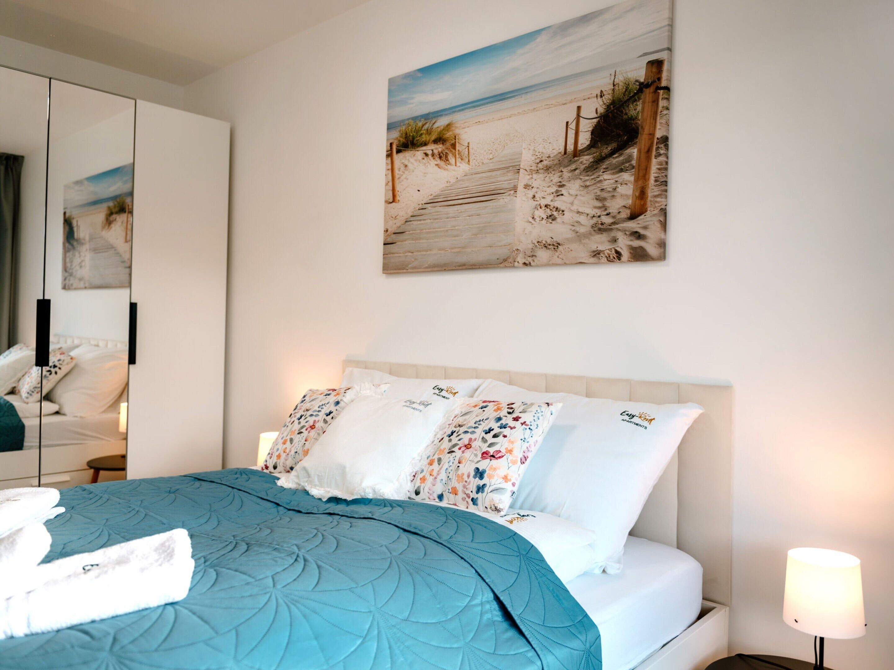 A bedroom with a bed, a nightstand, a lamp, and a beach-themed painting on the wall.