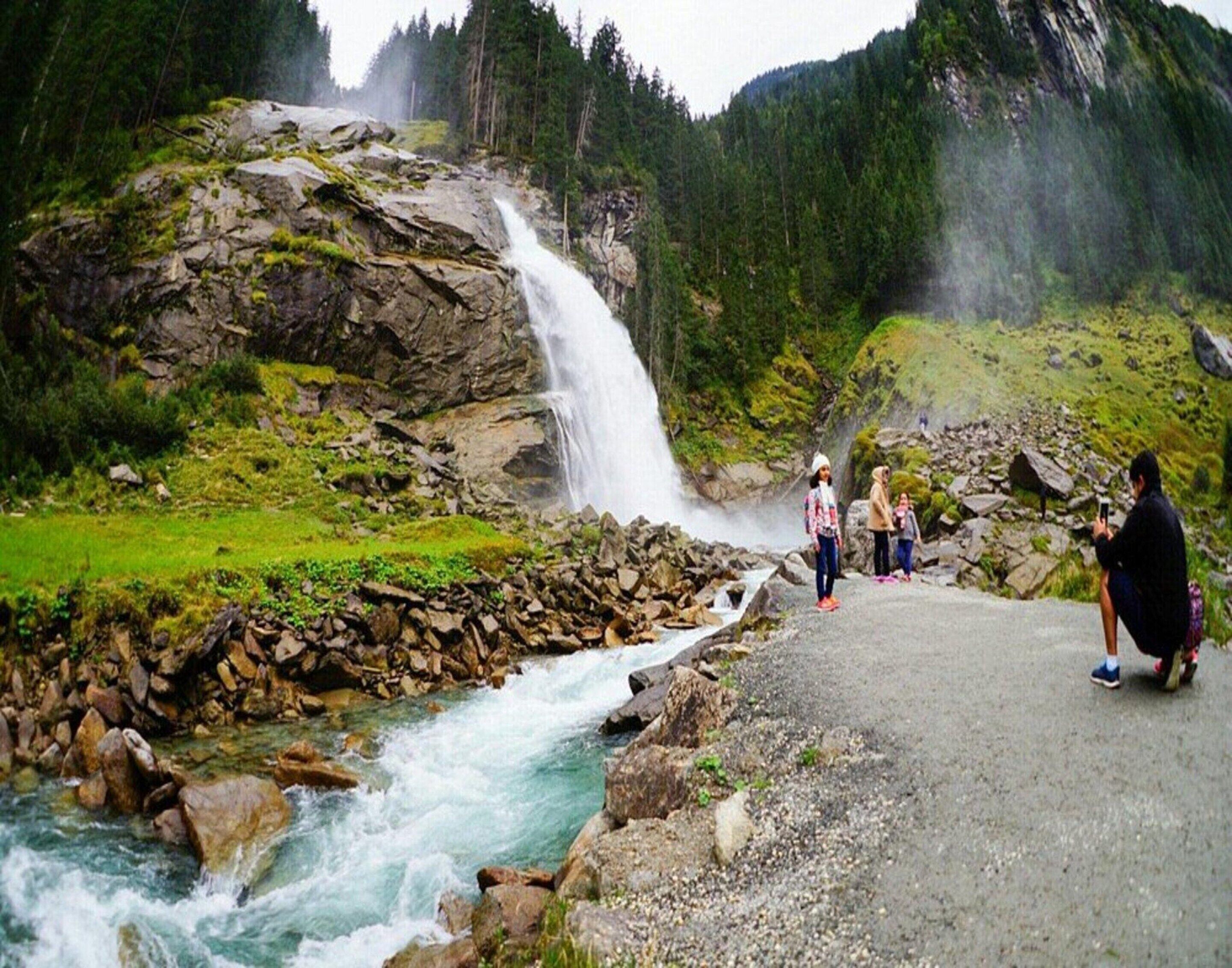 A waterfall cascading down a rocky cliff into a river, surrounded by lush greenery and a paved path with people observing.