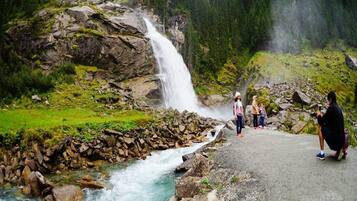 A waterfall cascading down a rocky cliff into a river, surrounded by lush greenery and a paved path with people observing.