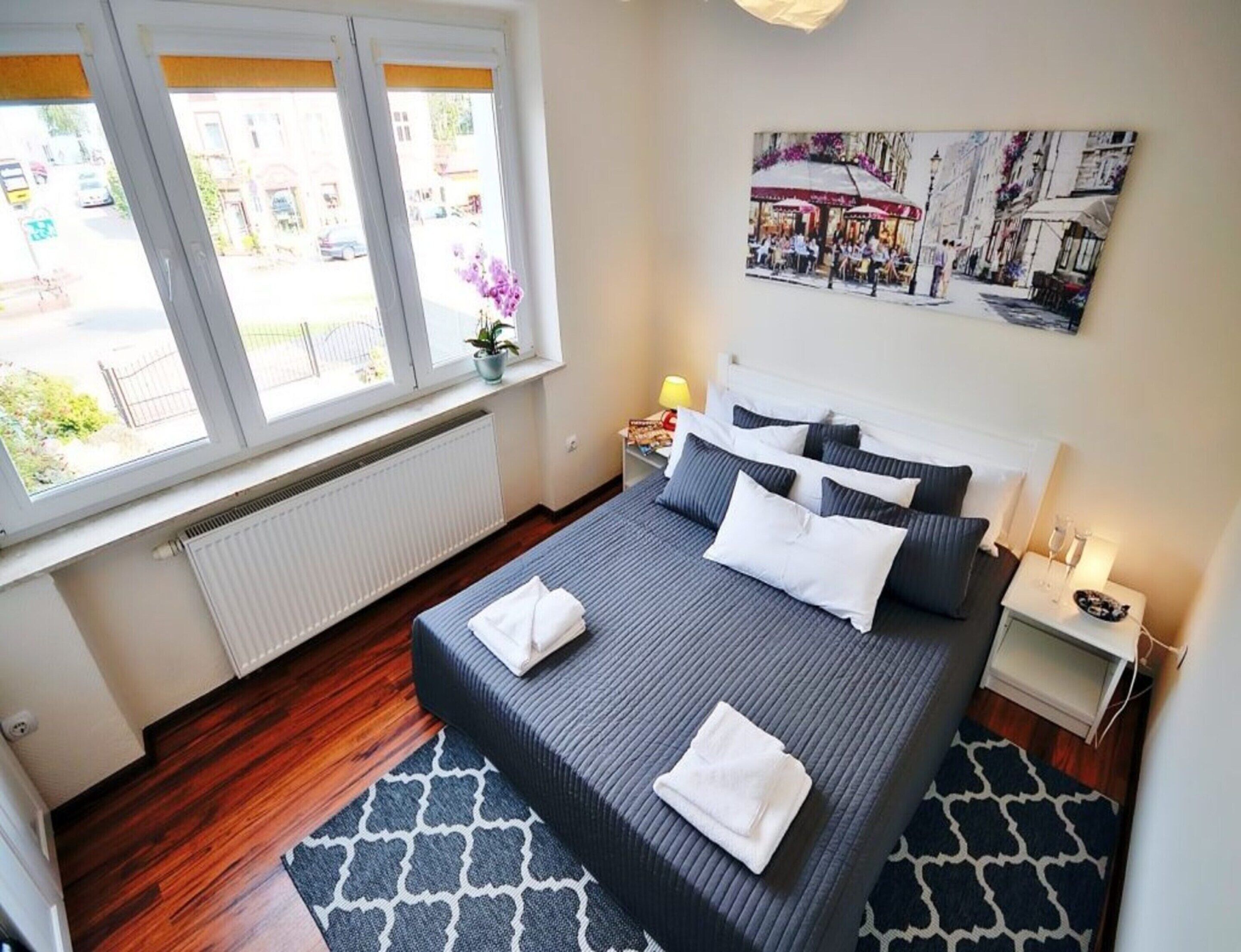 A neatly arranged bedroom with a bed, a nightstand, a window with a view of the street, and a wall-mounted picture.