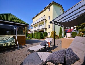 Un'area salotto all'aperto coperta, con poltrone e un tavolo, un edificio con balconi e una struttura con tetto verde.