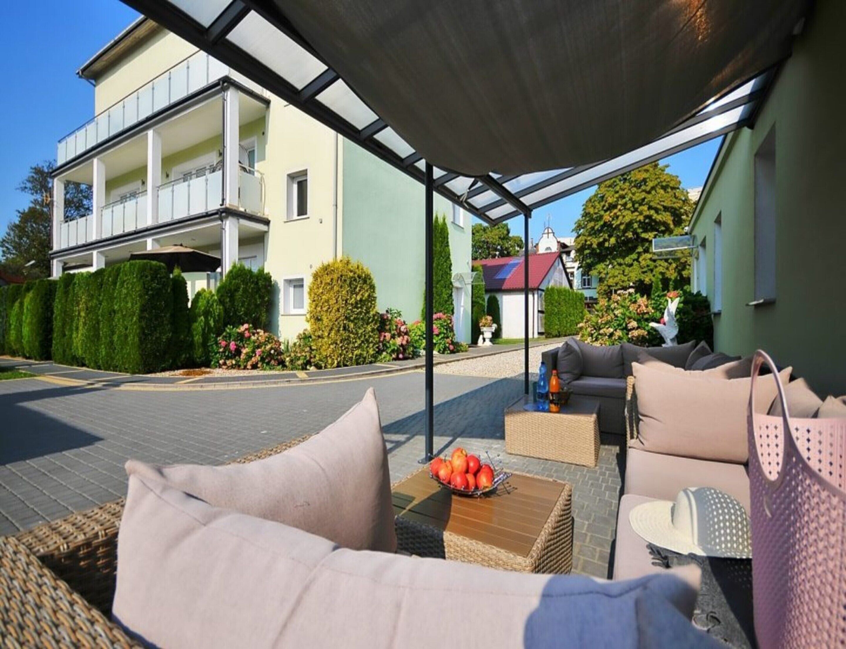 A covered outdoor seating area with wicker furniture, a table with a bowl of fruit, and a view of a building with balconies and greenery.
