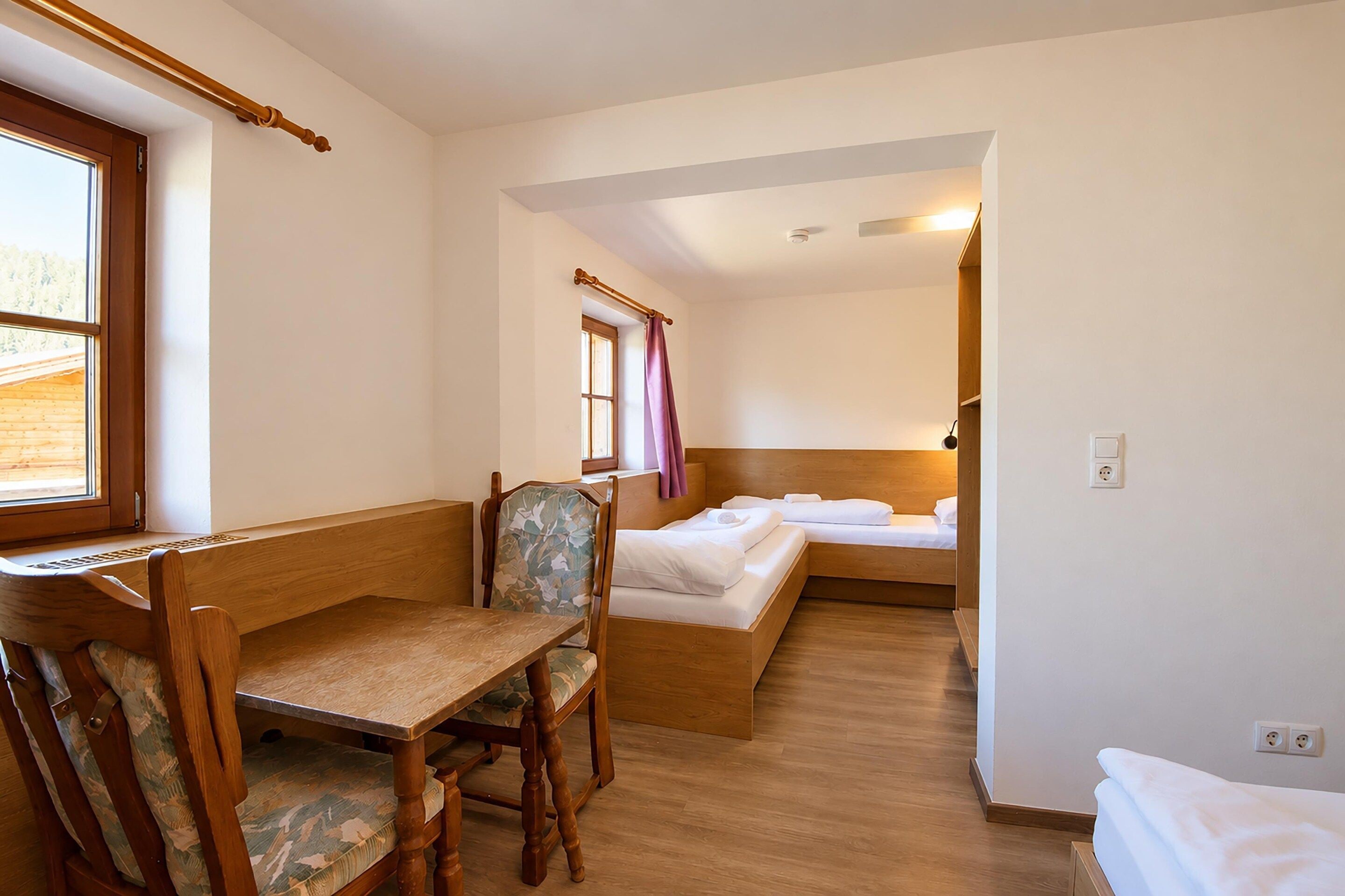 Una habitación con escritorio de madera, una silla, cama con ropa de cama blanca y una ventana con cortinas.