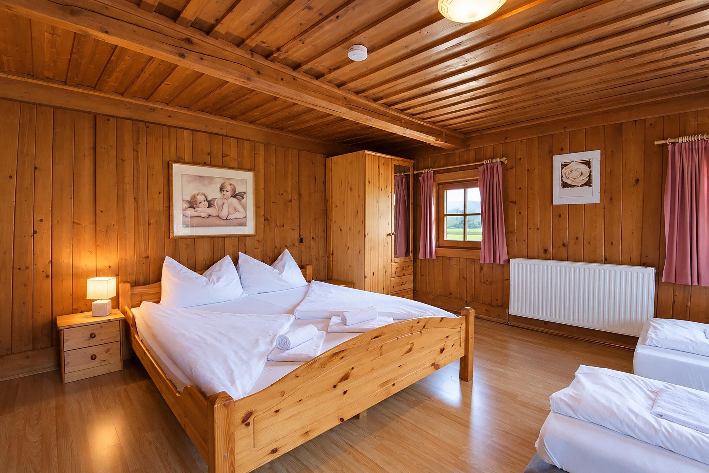 A wooden room with a bed, a nightstand, a window with curtains, a radiator, and a framed picture on the wall.