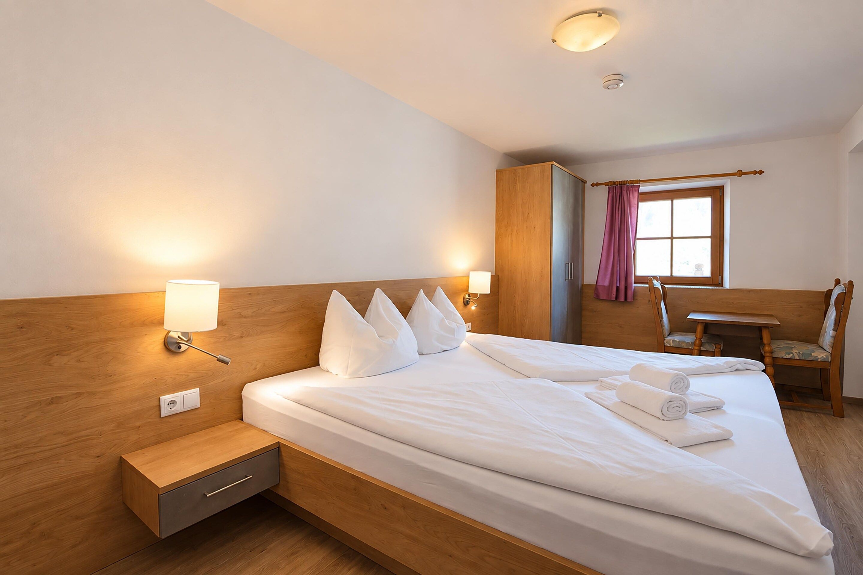 A hotel room with a large bed, bedside lamps, a wooden wardrobe, a desk, and a chair.