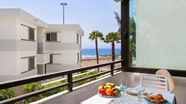 Un balcón con una mesa lista para el desayuno, con vista a una playa y un edificio blanco.