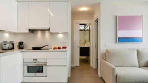 Una cocina moderna con armarios blancos, horno empotrado y cafetera. Hay un sofá y un cuadro en la pared.