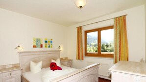 Ein Schlafzimmer mit einem hölzernen Bett, einem Nachttisch, einer Kommode, einem Fenster mit Vorhängen und Blick ins Freie.