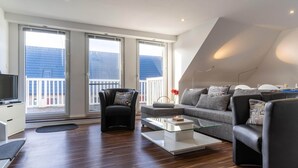 Ett modernt vardagsrum med en soffa, en fåtölj och ett soffbord i glas. Stora fönster ger utsikt över omgivningen.