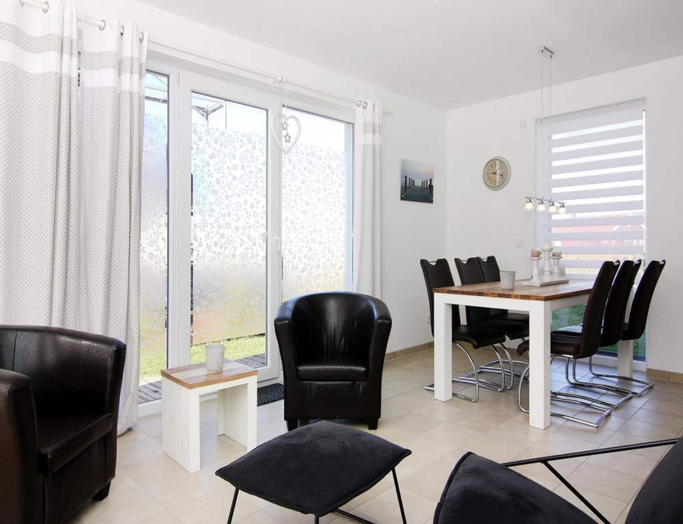 Un comedor moderno con una mesa blanca y sillas negras, ventanas amplias con cortinas, y vistas a zonas verdes al exterior.