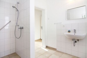 Une salle de bain avec des murs recouverts de carreaux blancs, une douche avec un pommeau de douche amovible, un lavabo avec un miroir au-dessus et une petite étagère contenant des articles de toilette.