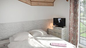 Ein Schlafzimmer mit weißem Ziegel-Kopfstück, einem Bett mit weißer Bettwäsche und Kissen mit Herzmuster, einem Fernseher auf einem Nachttisch und einem Fenster mit floralen Vorhängen.